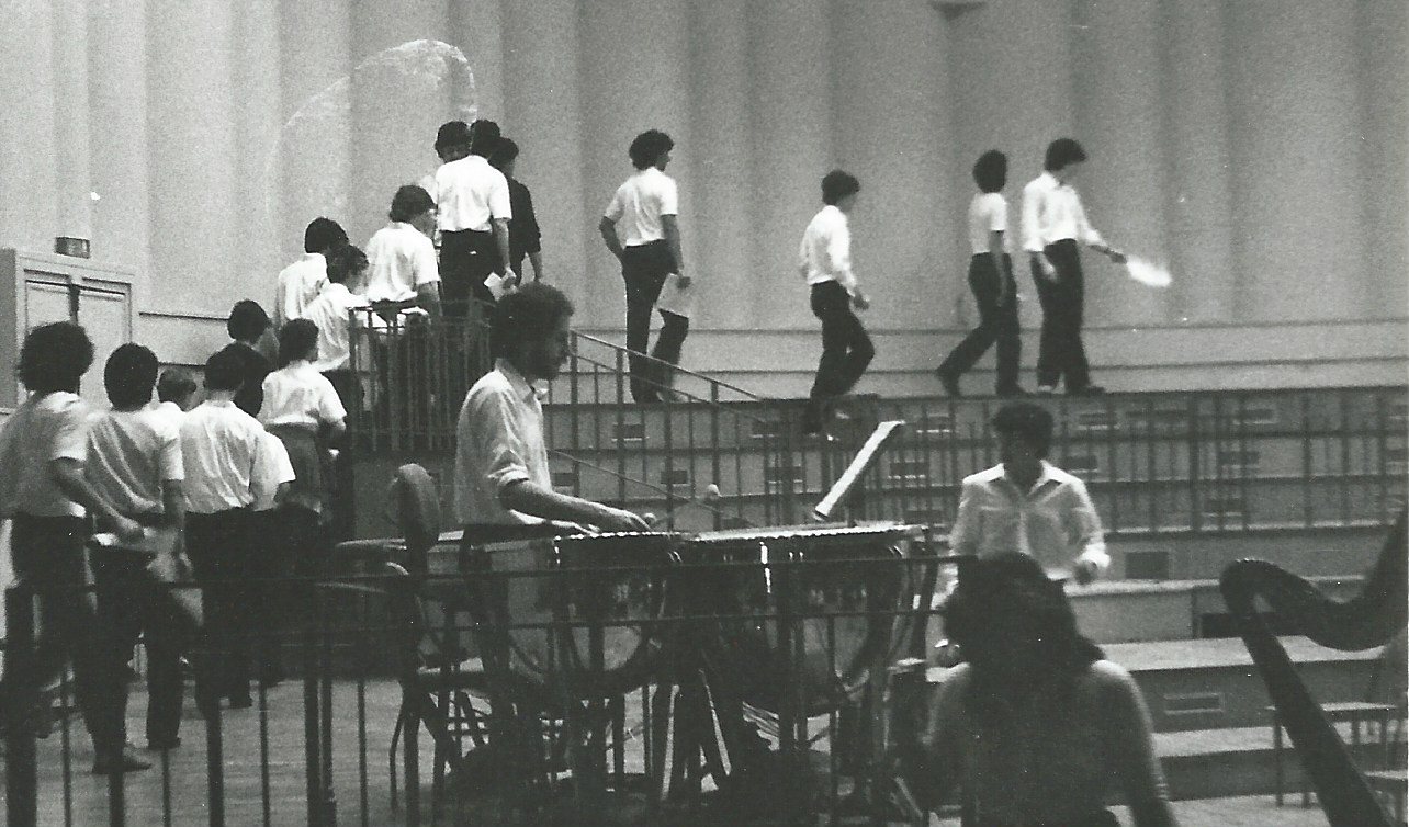 Timpani in primo piano, coro che sale sul palco